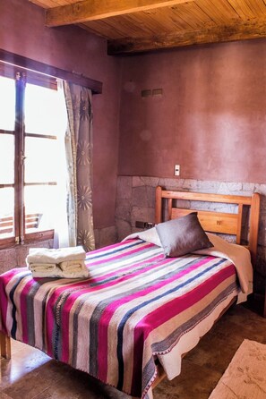 Quarto com cabeceira de madeira, cama com manta listrada, mesa de cabeceira com toalhas e janela com cortinas.