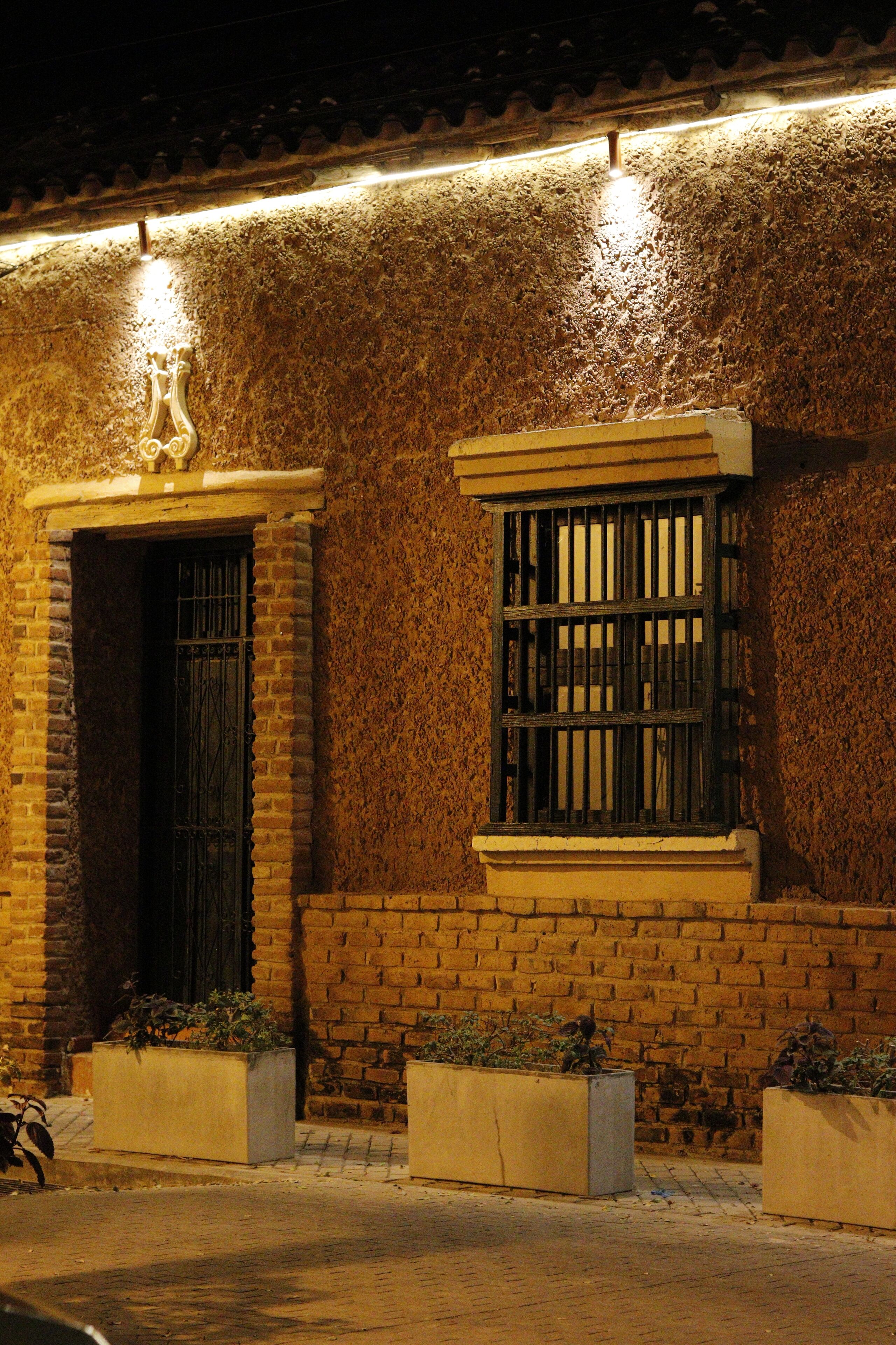 A building with a brick facade, a window with bars, and a door with a metal gate. There are planters with plants in front of the door.