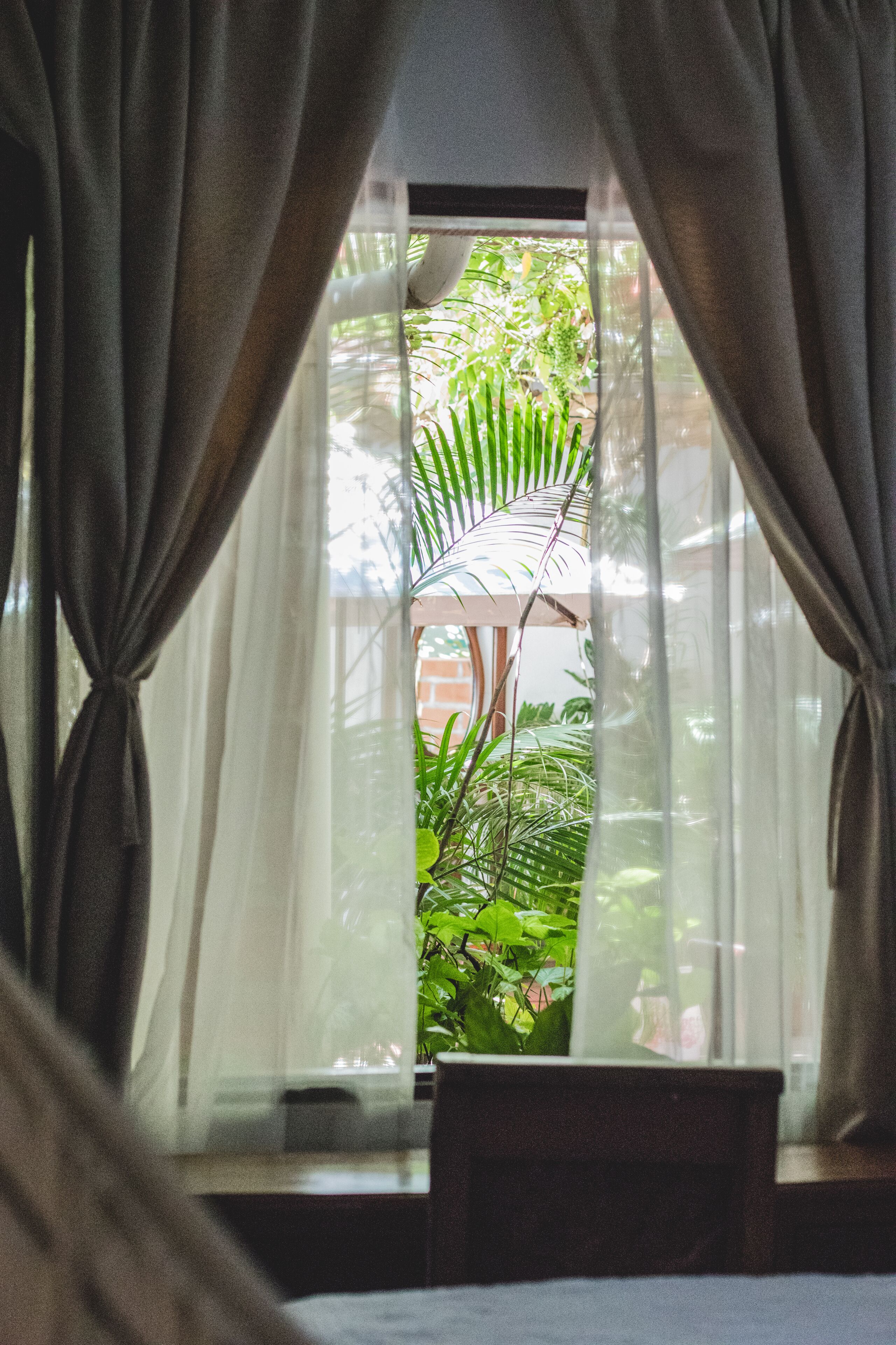 A room with a window covered by sheer curtains, a wooden bench, and a view of greenery outside.