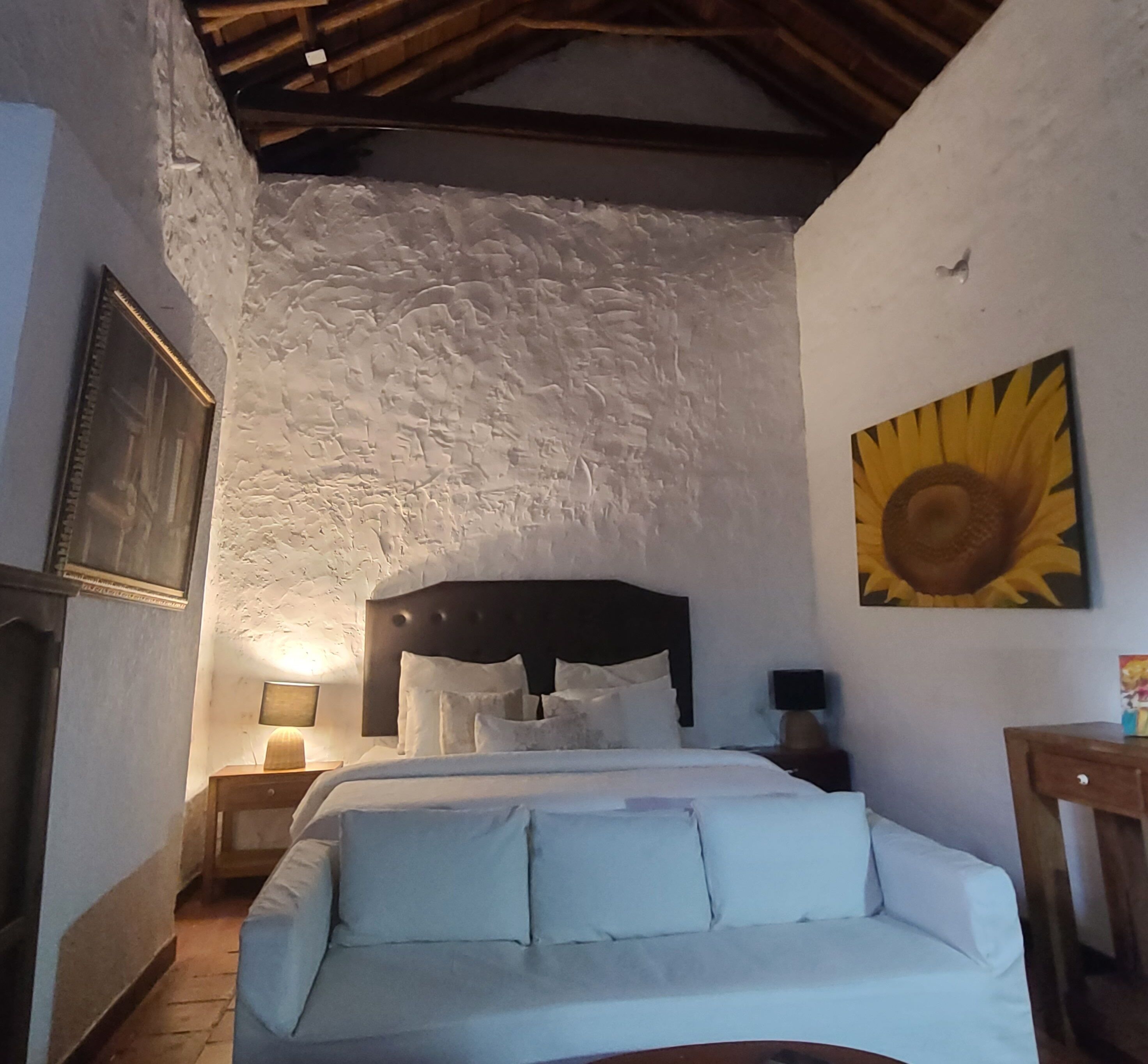 Una camera da letto con parete bianca strutturata, soffitto in legno, un letto con biancheria da letto bianca, un comodino con una lampada e un quadro incorniciato appeso al muro.