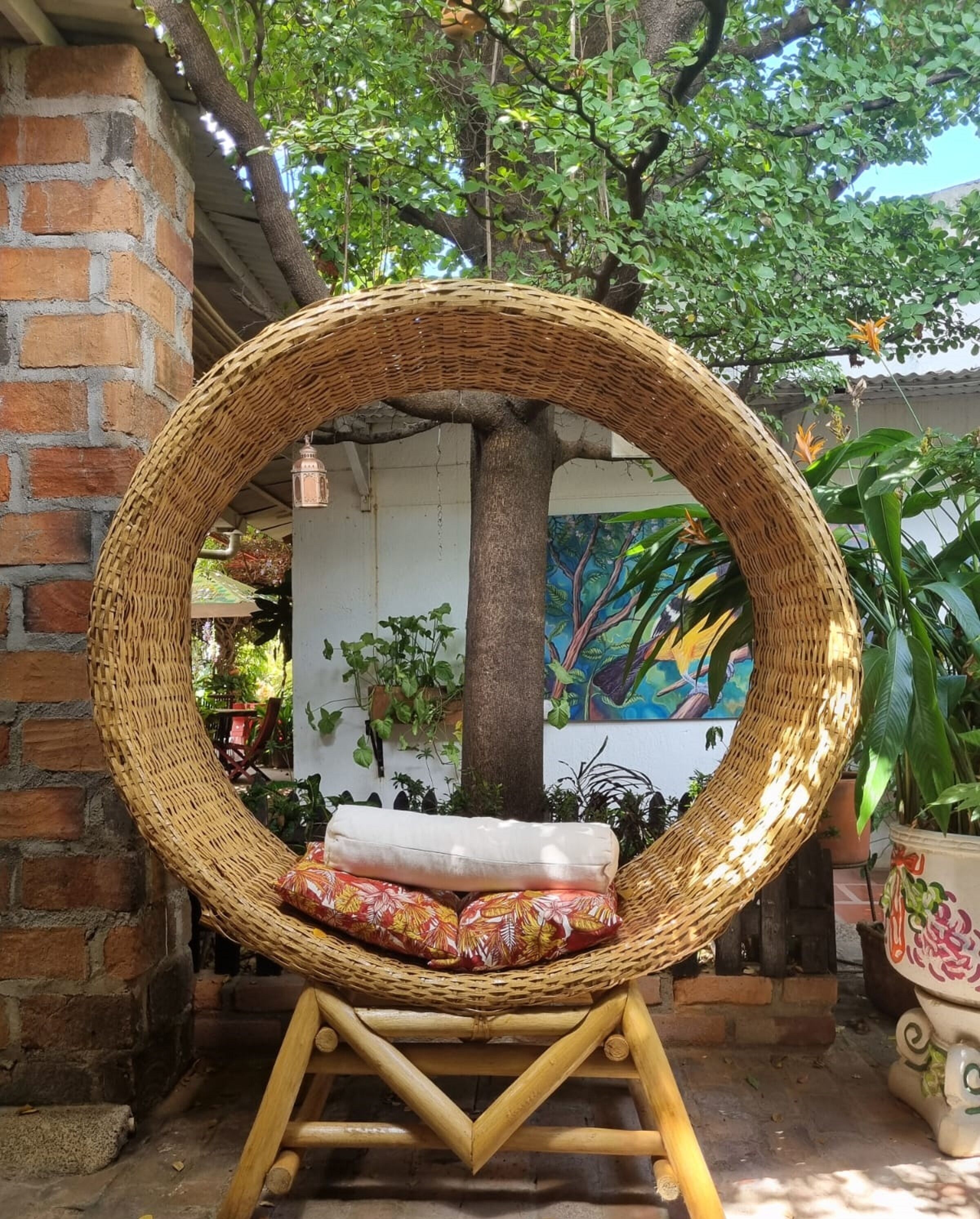 Uma cadeira de vime com almofada, colocada ao ar livre, com uma árvore e plantas ao fundo.