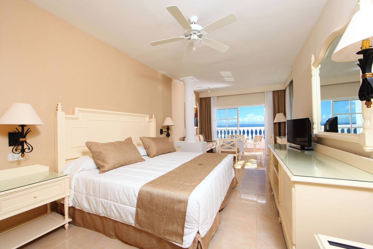 A hotel room with a large bed, a ceiling fan, a TV, and a view of the ocean.