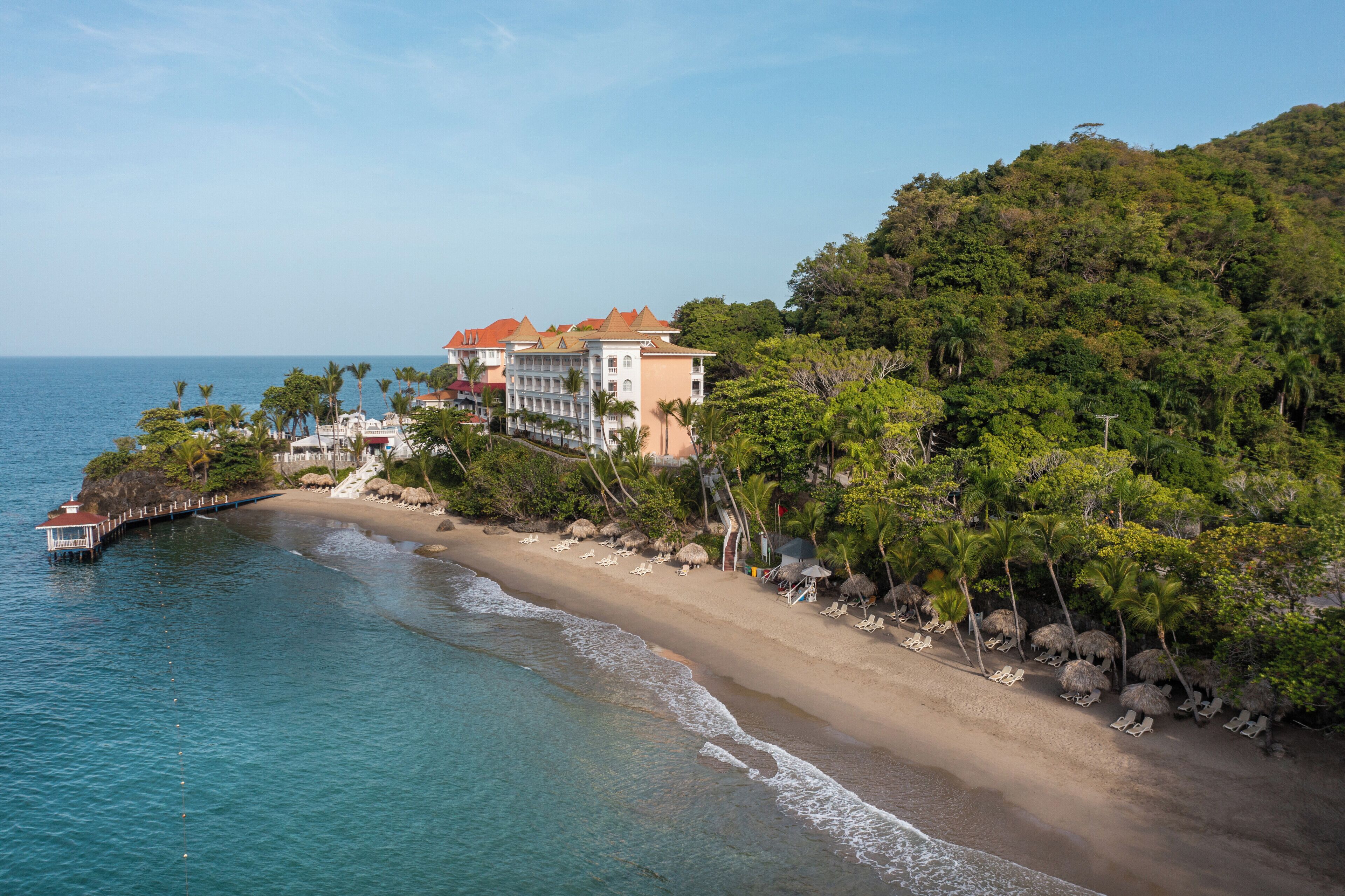 Un hotel frente al mar con un muelle, playa y exuberante vegetación.