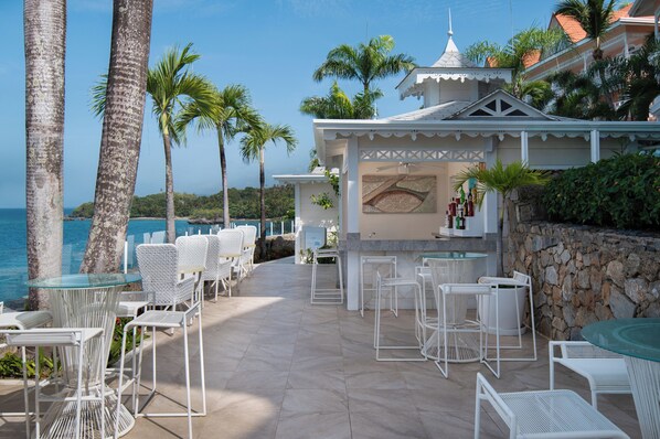 Une terrasse avec des chaises et des tables en osier blanc, un mur de pierre et une vue sur l’océan.