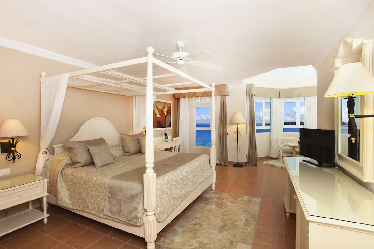 Un lit à baldaquin, un bureau avec une lampe et une vue sur la mer grâce à de grandes fenêtres.