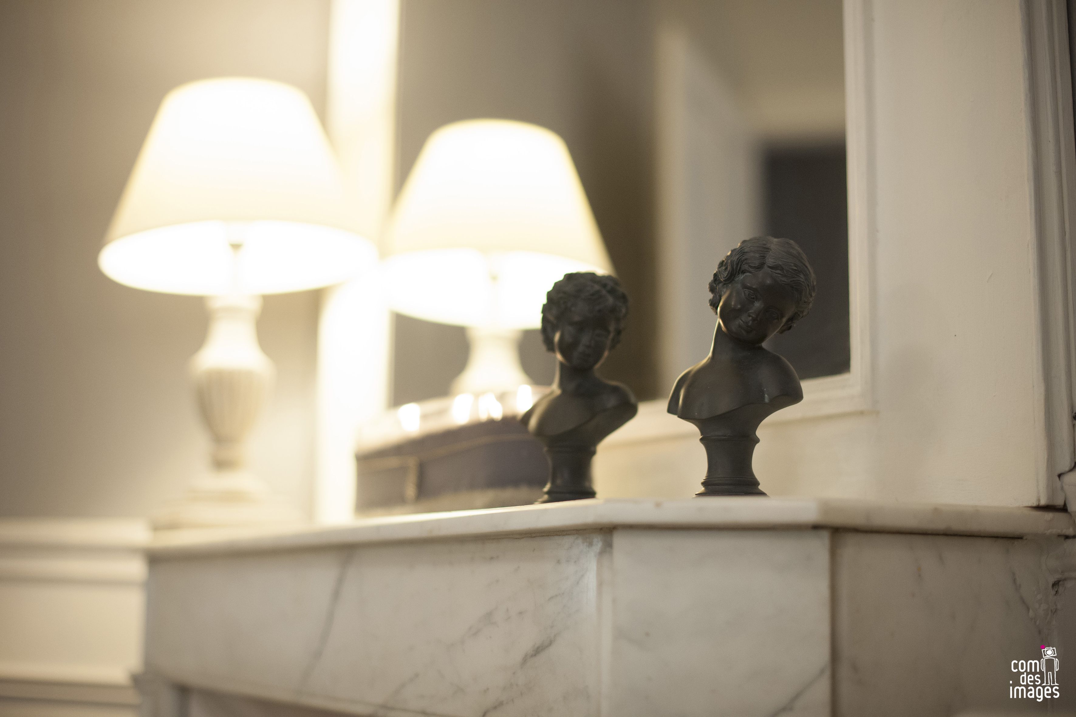 A marble fireplace with two bust sculptures and two lit lamps.