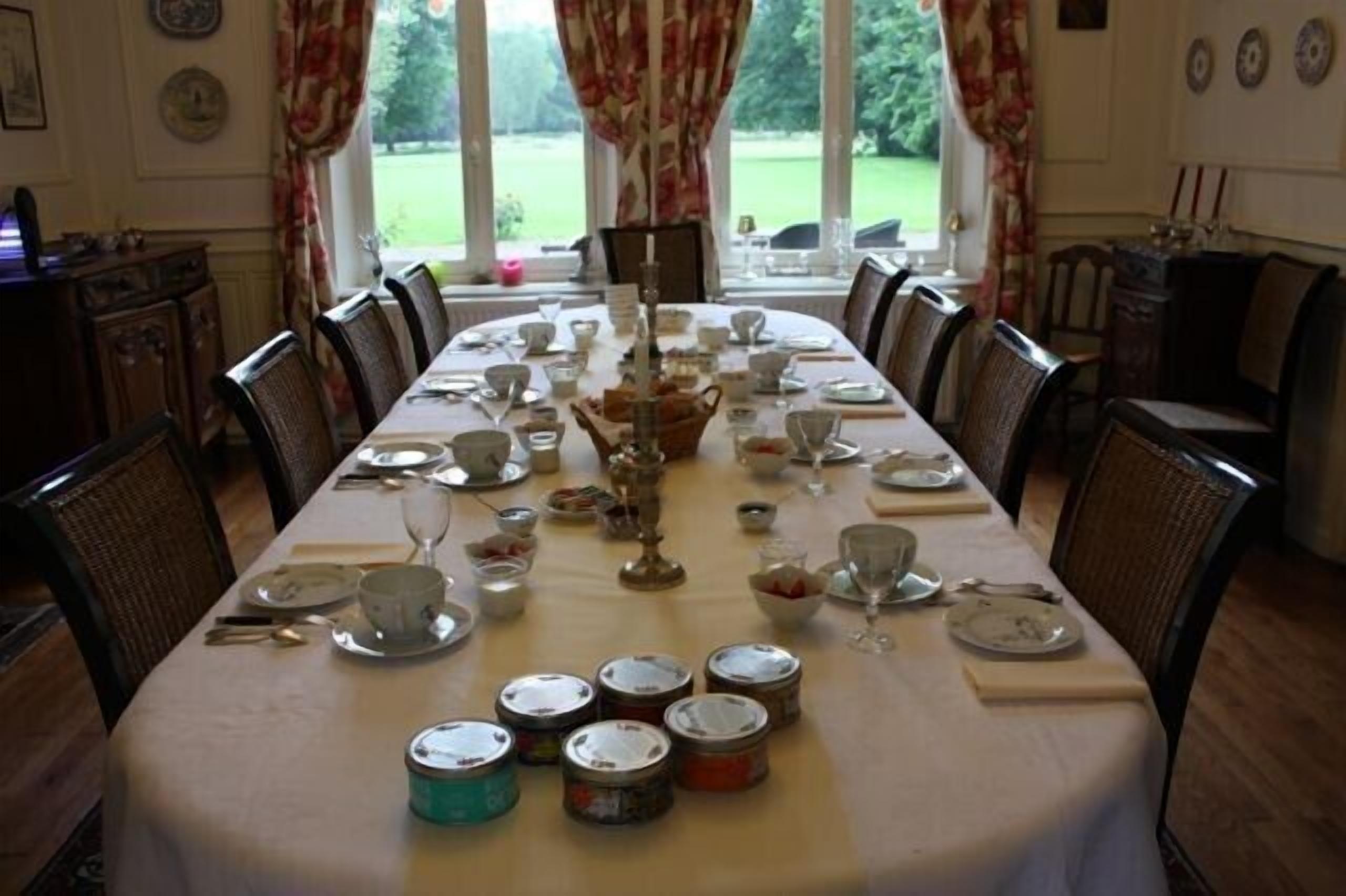 A dining room with a long table set for a meal, surrounded by chairs, with a view of a garden through large windows.