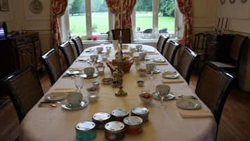 A dining room with a long table set for a meal, surrounded by chairs, with a view of a garden through large windows.