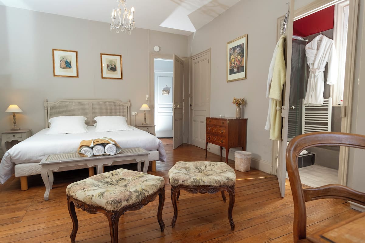 Une chambre à coucher comprenant un lit, deux fauteuils rembourrés, une commode en bois et une porte rouge donnant accès à une salle de bain.