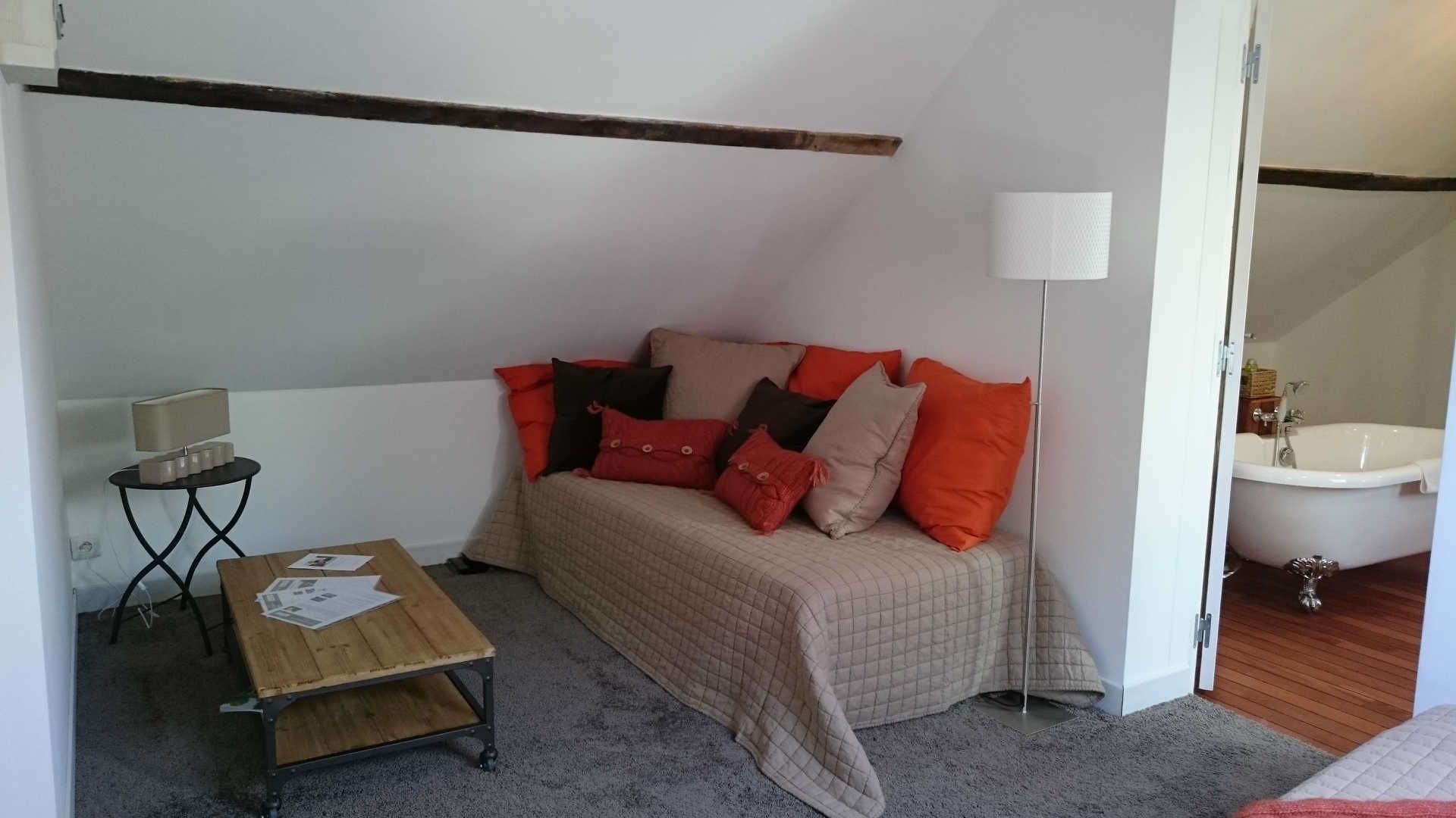 Una habitación pequeña y acogedora con una cama individual, una mesa de madera con papeles, una lámpara y un baño visible a través de una puerta abierta.