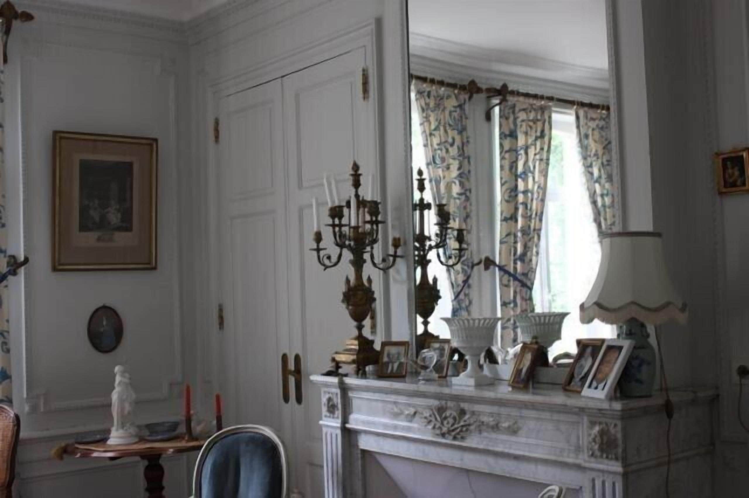A room with a white fireplace, a mirror, a chandelier, and a window with patterned curtains.