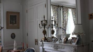 A room with a white fireplace, a mirror, a chandelier, and a window with patterned curtains.