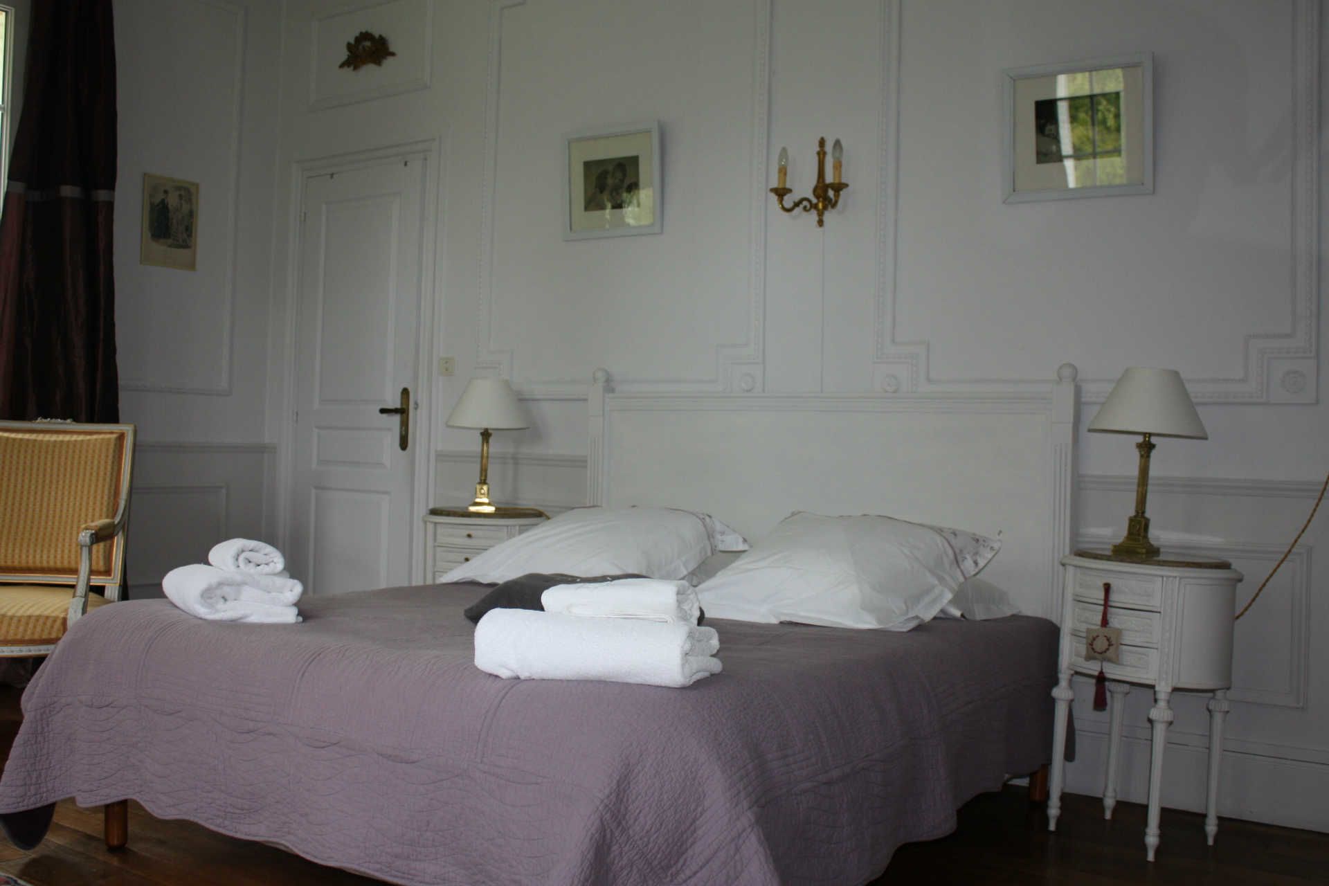 A neatly made bed with white linens and towels, a bedside table with a lamp, and a chair with folded towels.