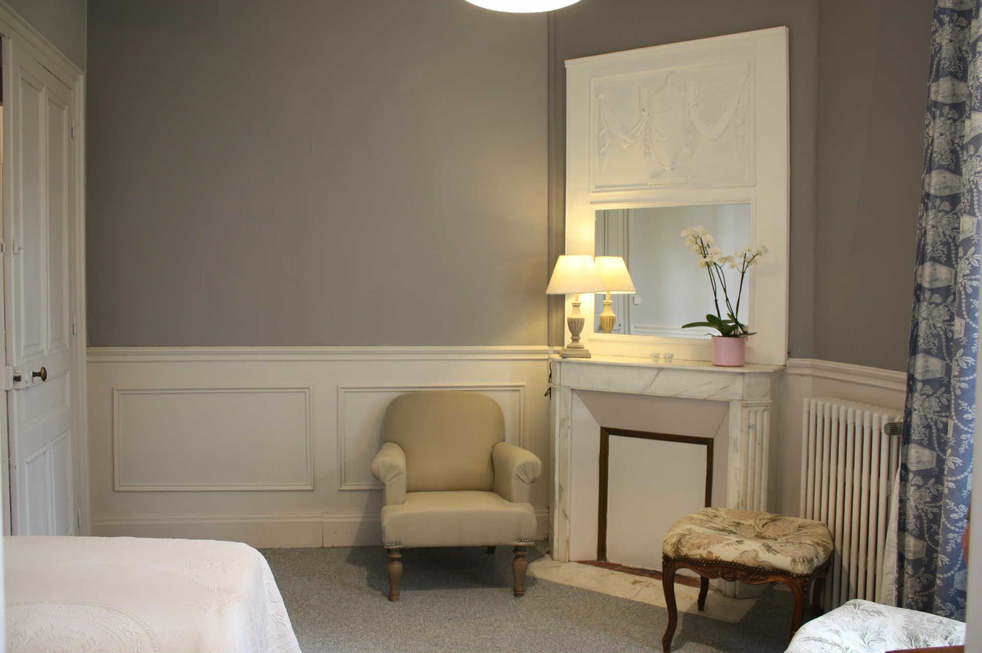 Ein Zimmer mit einem beigefarbenen Sessel, einem kleinen Hocker, einem weißen Bett, einem Kamin und einem Spiegel mit Lampe.