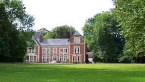 Een groot, historisch gebouw met een symmetrische gevel, omringd door een verzorgd gazon en weelderige bomen.