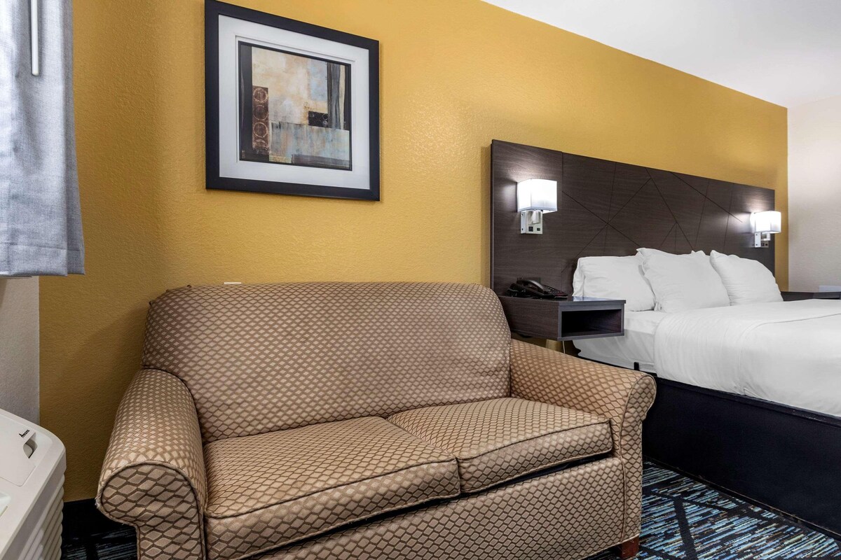 Habitación de hotel con un sofá estampado, una cama con ropa blanca, una obra de arte enmarcada en la pared y una mesita de noche con una lámpara.