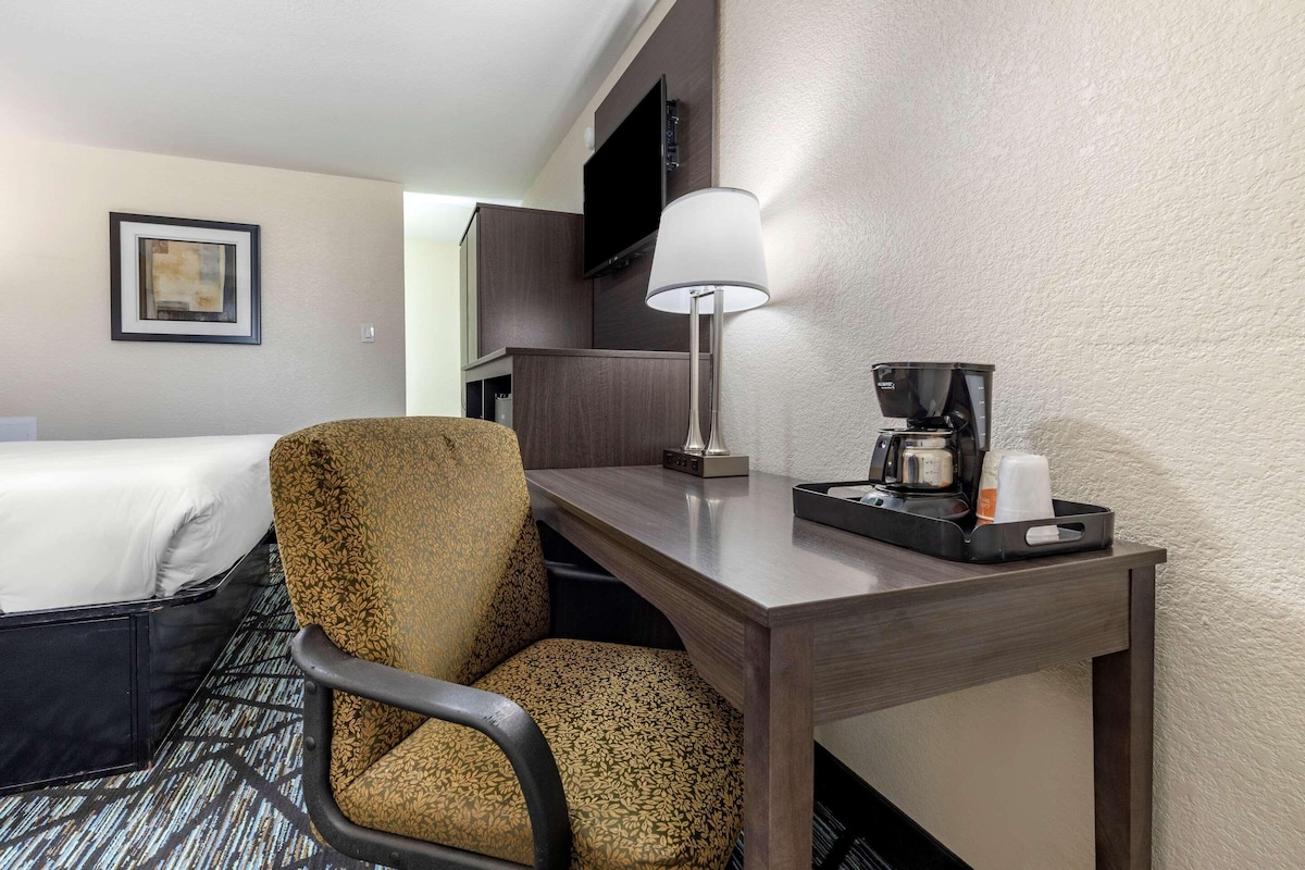 Habitación de hotel con cama, escritorio con lámpara, cafetera y un cuadro en la pared.
