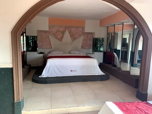 A hotel room with a large bed, a nightstand, a mirror, and a view of the outside.