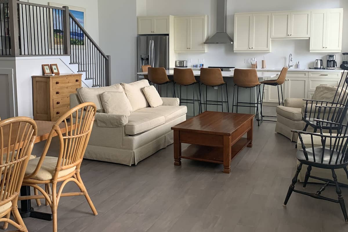 A modern living room with a sofa, chairs, and a coffee table. There is a kitchen area with cabinets and appliances. A staircase with a railing is visible in the background.