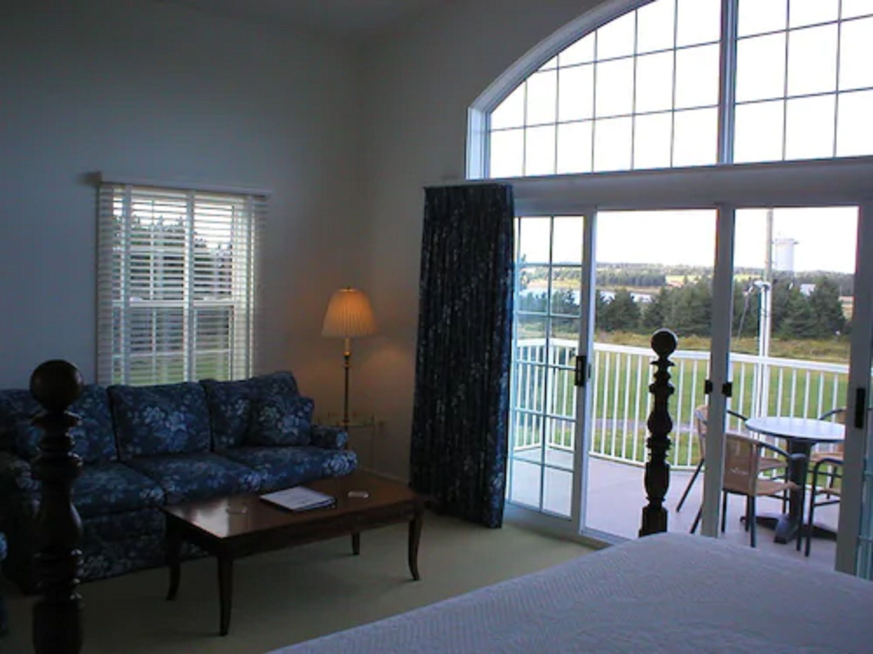 A room with a bed, a sofa, a coffee table, a lamp, and a balcony with a view of the outdoors.