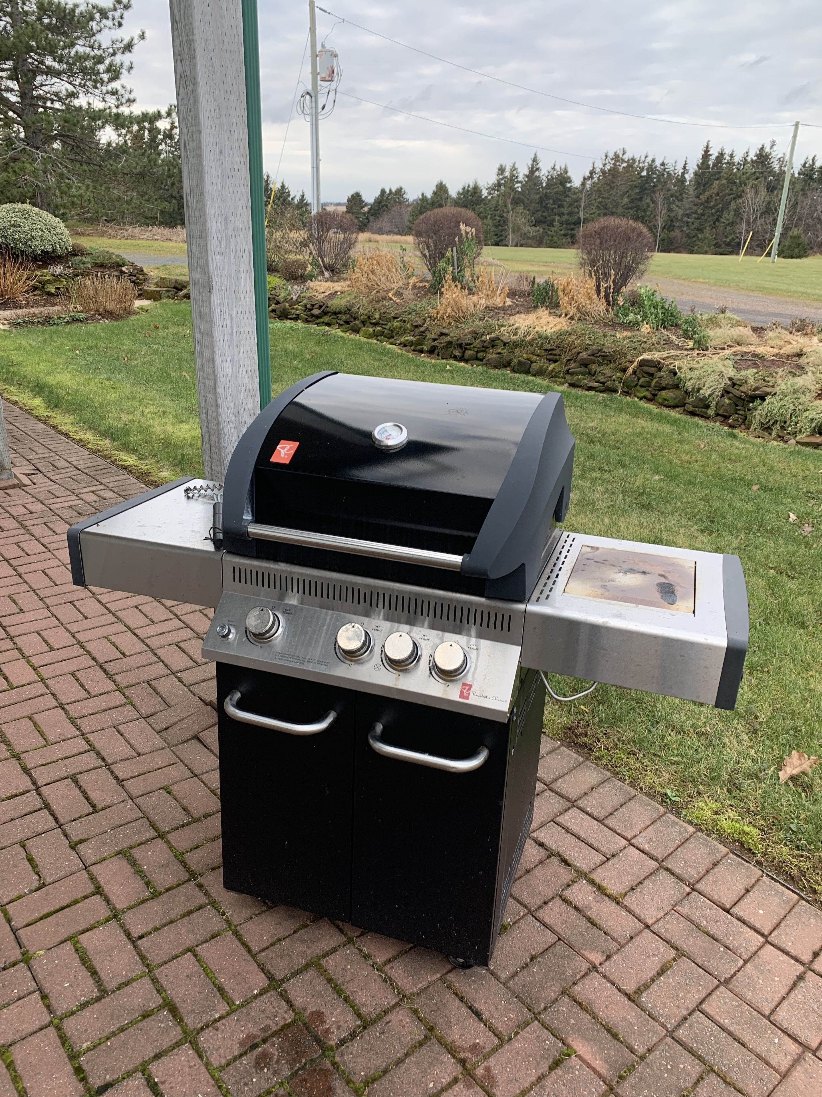 A portable gas grill with four burners and a side burner, placed on a brick patio.