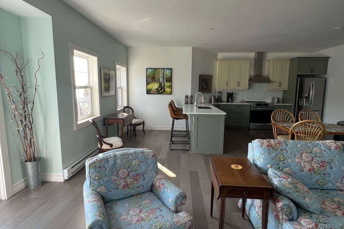 A spacious living room with floral-patterned sofas, a dining area with wooden chairs, and a kitchen with light-colored cabinets.
