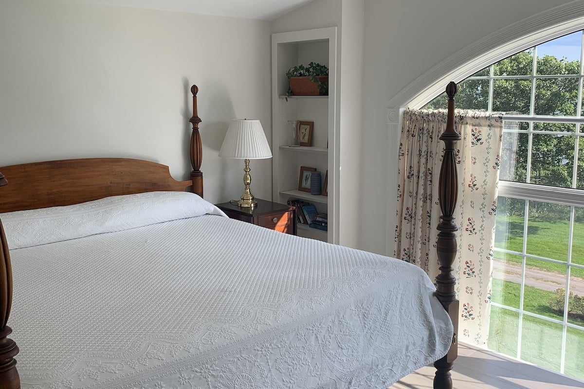A bedroom with a wooden bed, a nightstand with a lamp, a shelf with decorative items, and a window with curtains.