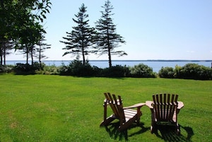 Two Adirondack chairs on a grassy lawn with a view of the water and trees.