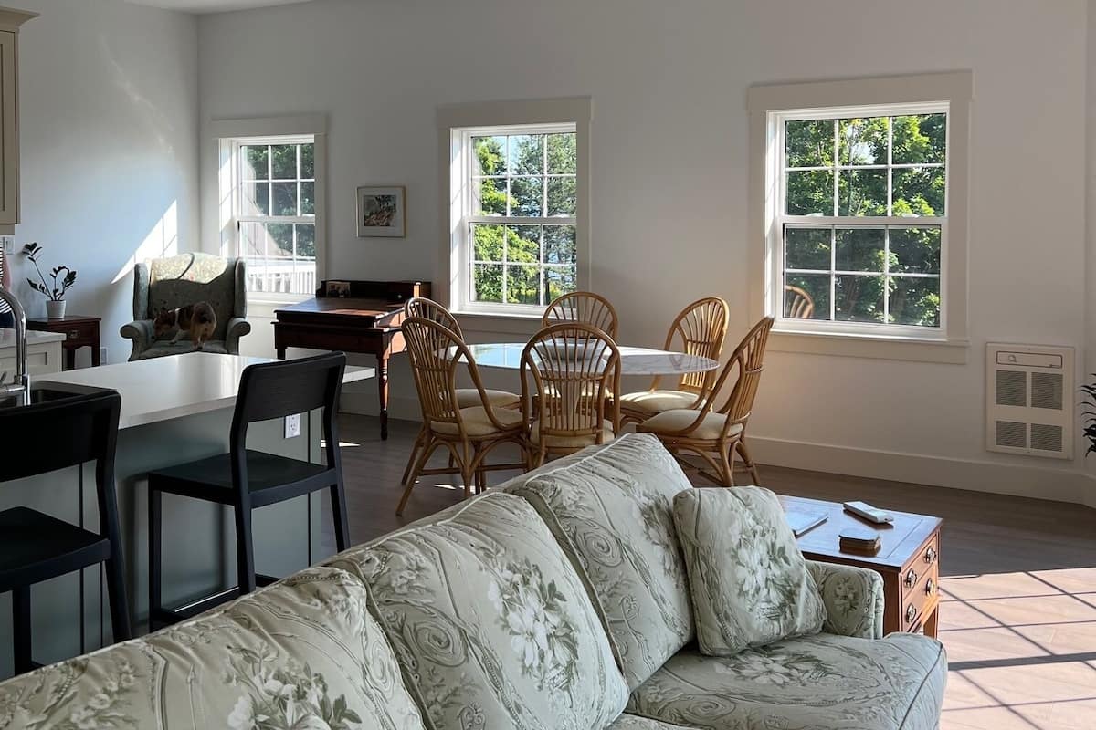 A spacious living room with a large floral sofa, a dining area with wicker chairs, and large windows providing natural light.