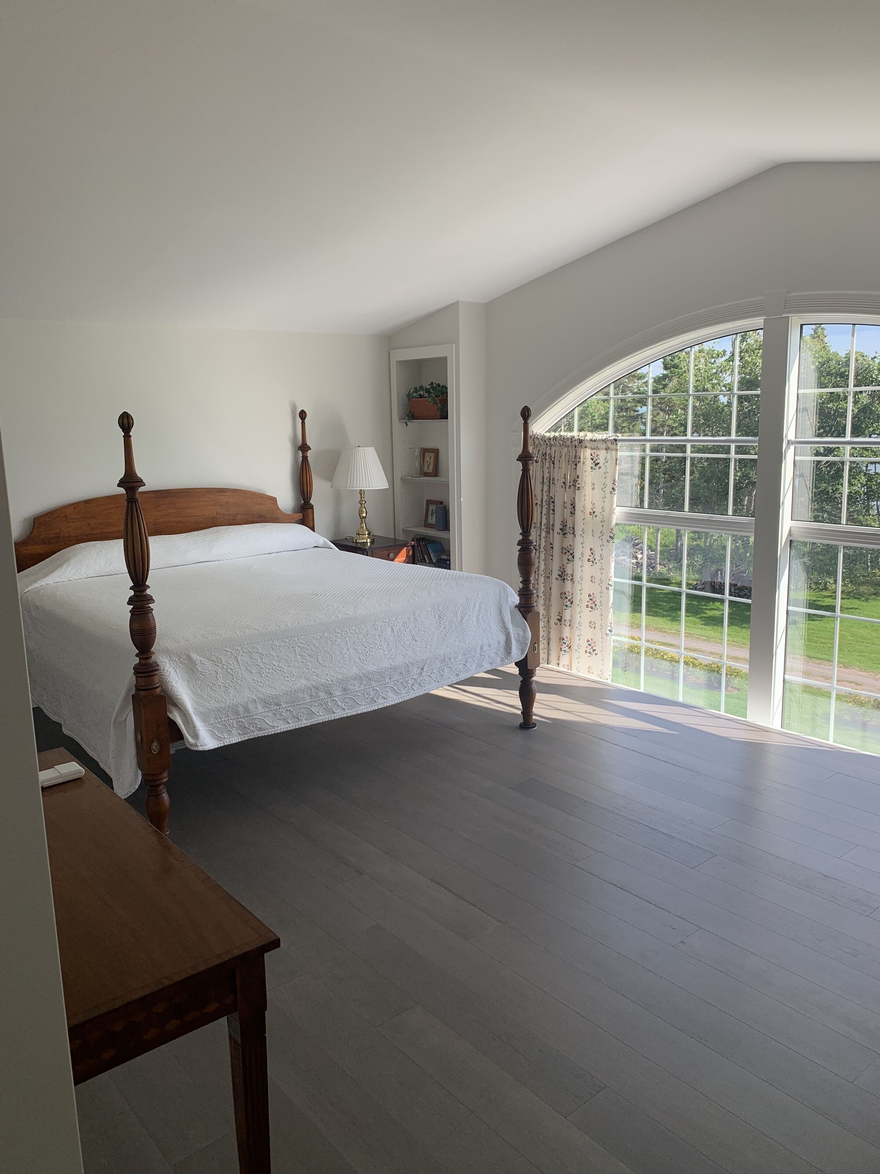 A bedroom with a large bed, a nightstand, a lamp, a wooden table, and a view of greenery through a large window.