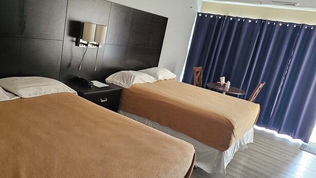 A hotel room with two beds, a nightstand, a lamp, a chair, and a table with a curtain. - Town N Country Motor Inn (Leamington)