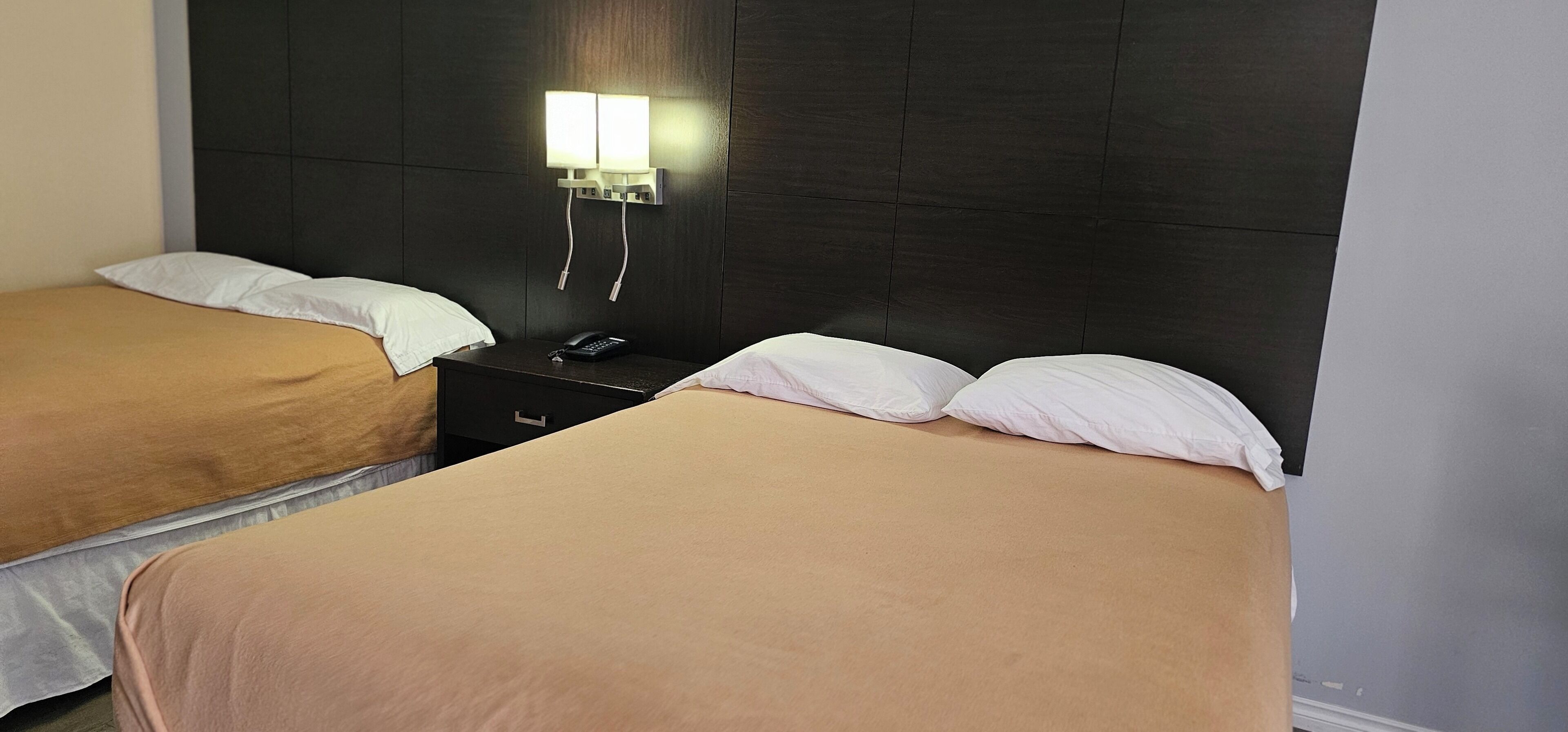 Quarto de hotel com duas camas, cada uma com colcha bege e almofadas brancas. Há uma mesa de cabeceira com um telefone e um abajur entre as camas.