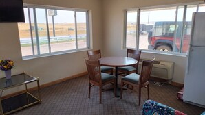 A small room with a round table and four chairs, a television on the wall, and large windows with a view of a parking lot and a red vehicle.
