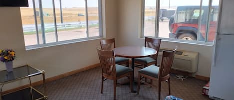 Una habitación pequeña con una mesa redonda y cuatro sillas, un televisor en la pared y ventanas amplias con vista a un estacionamiento y un vehículo rojo.
