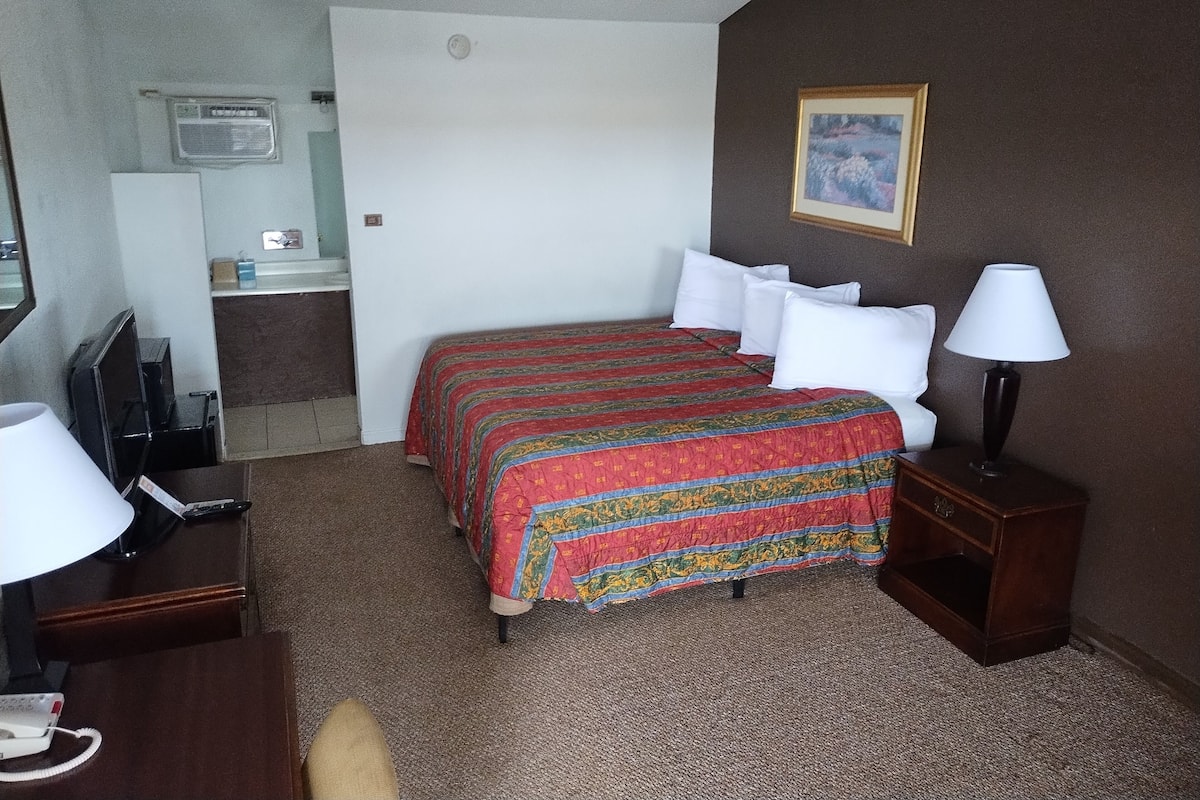 A hotel room with a bed, a desk with a lamp, a chair, a TV, and a bathroom visible through an open door.