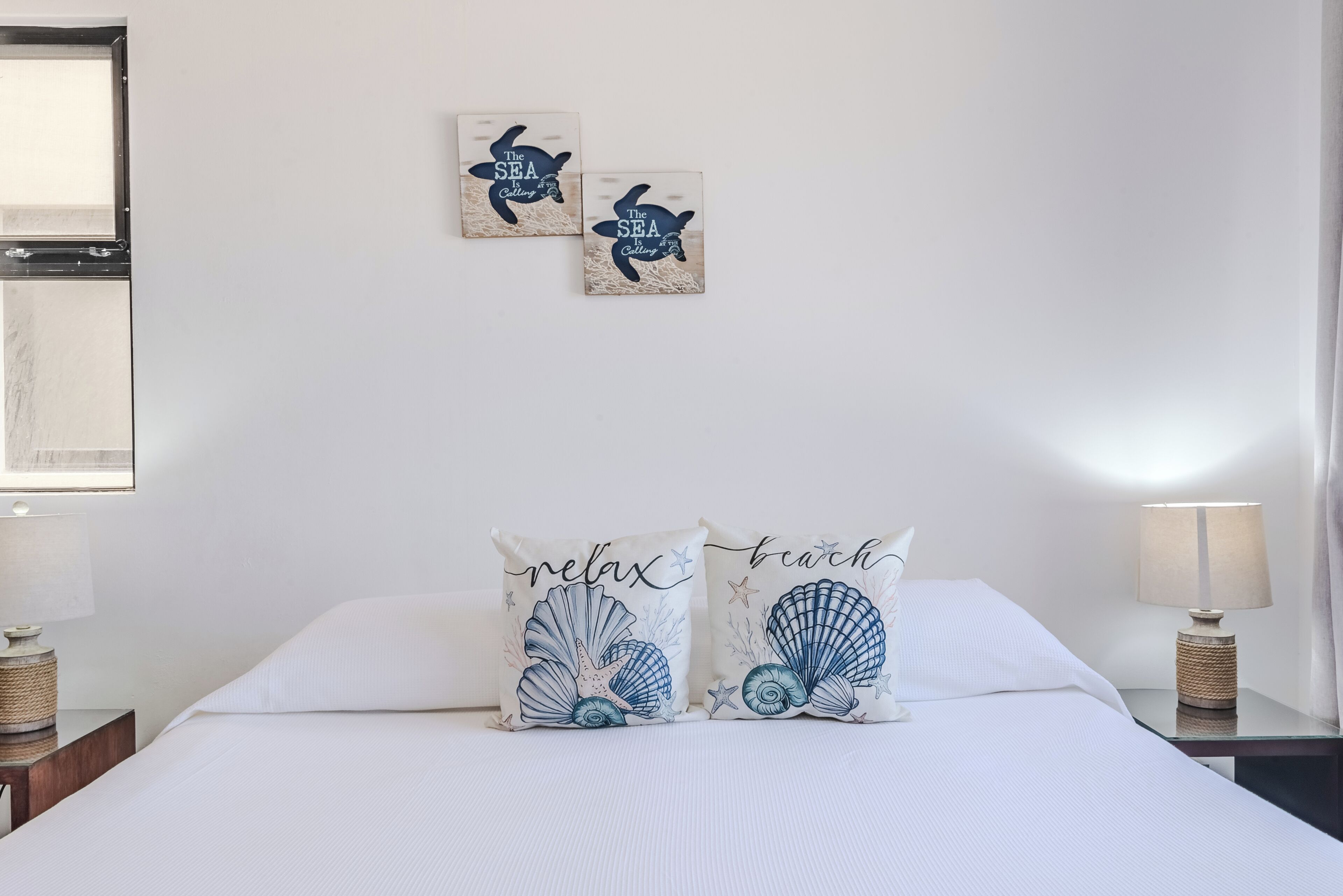 A bedroom with a bed, two pillows with sea-themed designs, a nightstand with a lamp, and a wall-mounted picture.