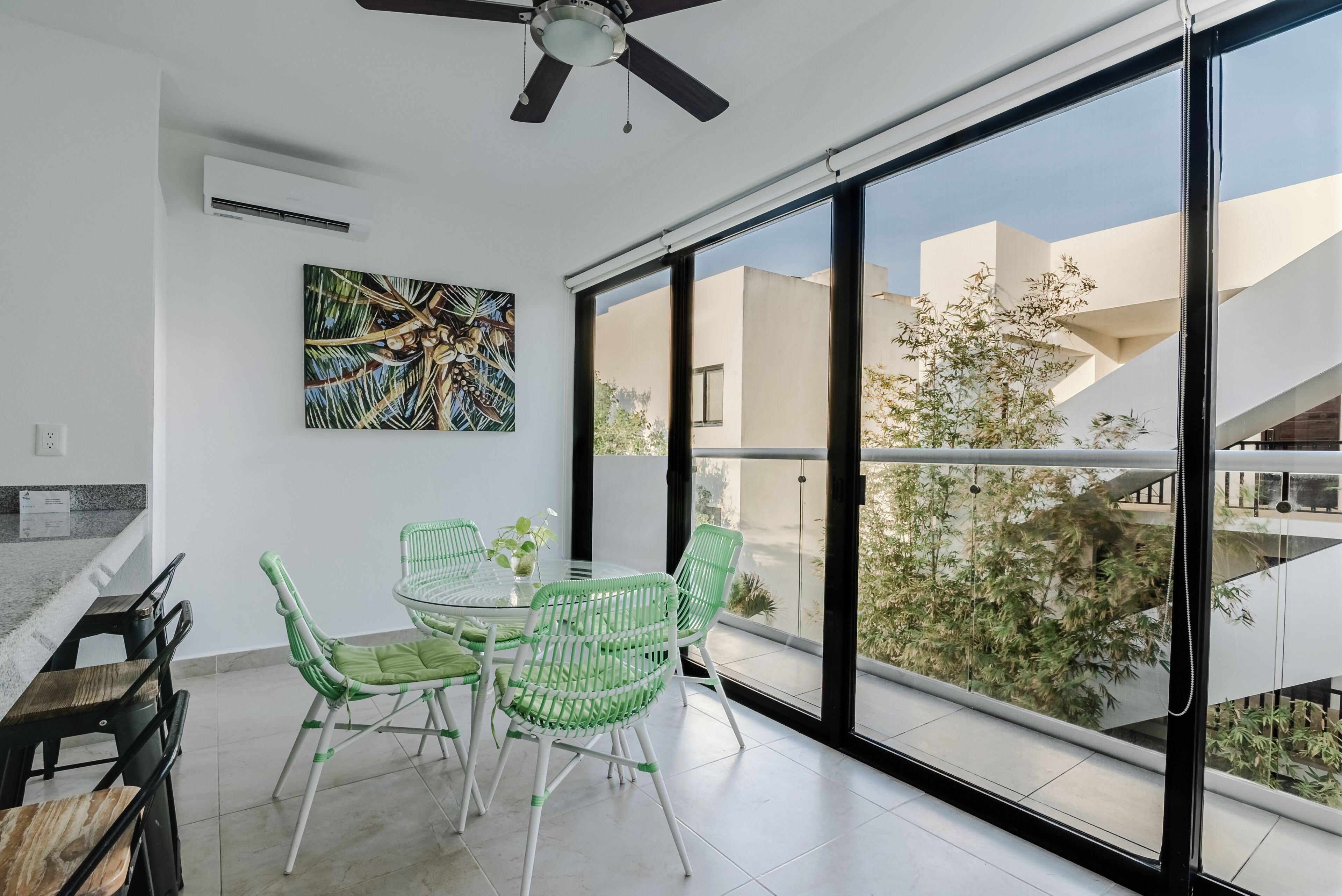 A modern dining area with a glass table and green chairs, a ceiling fan, and a large window with a view of buildings and greenery.