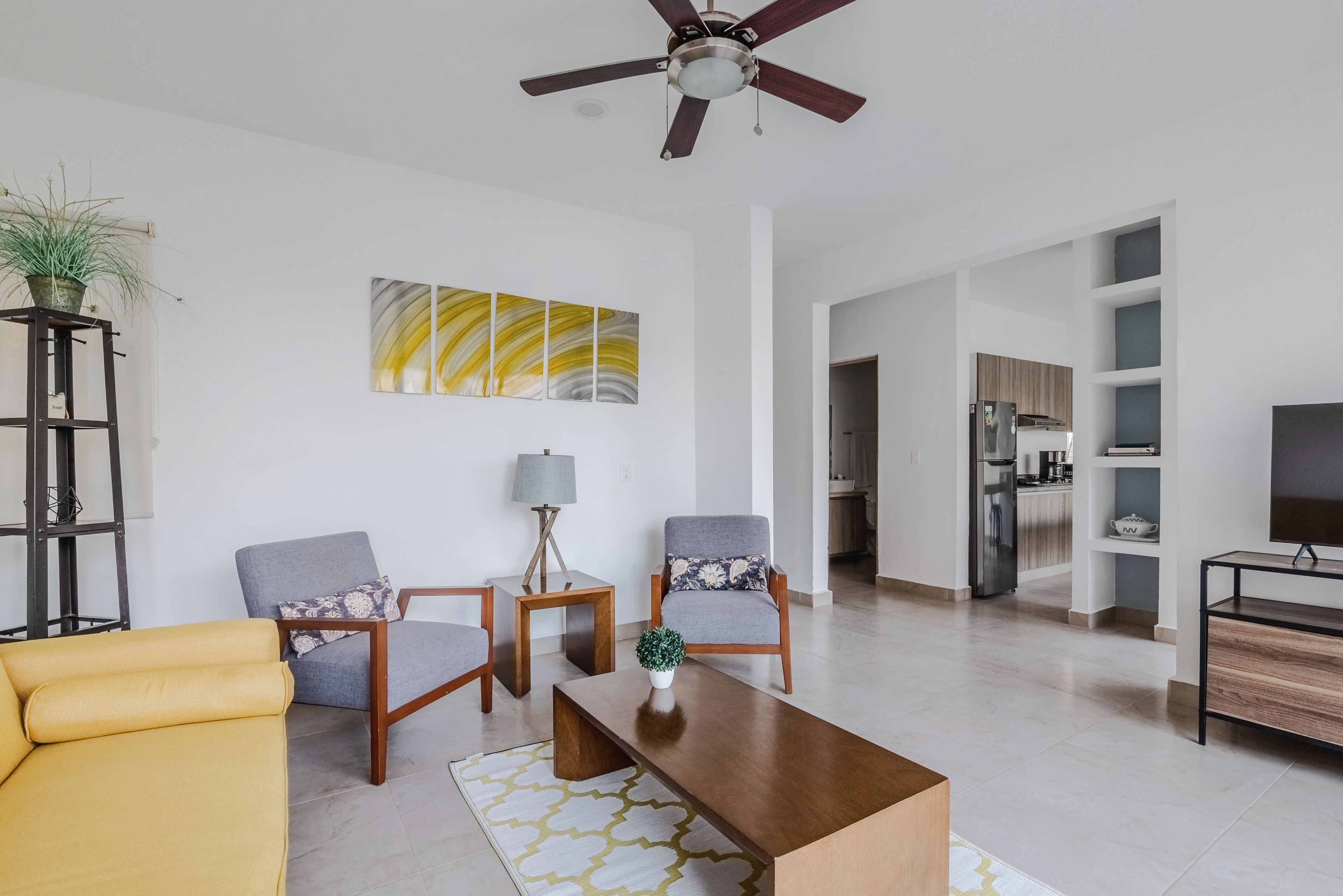 A modern living room with a sofa, chairs, a coffee table, and a ceiling fan. A kitchen is visible in the background.