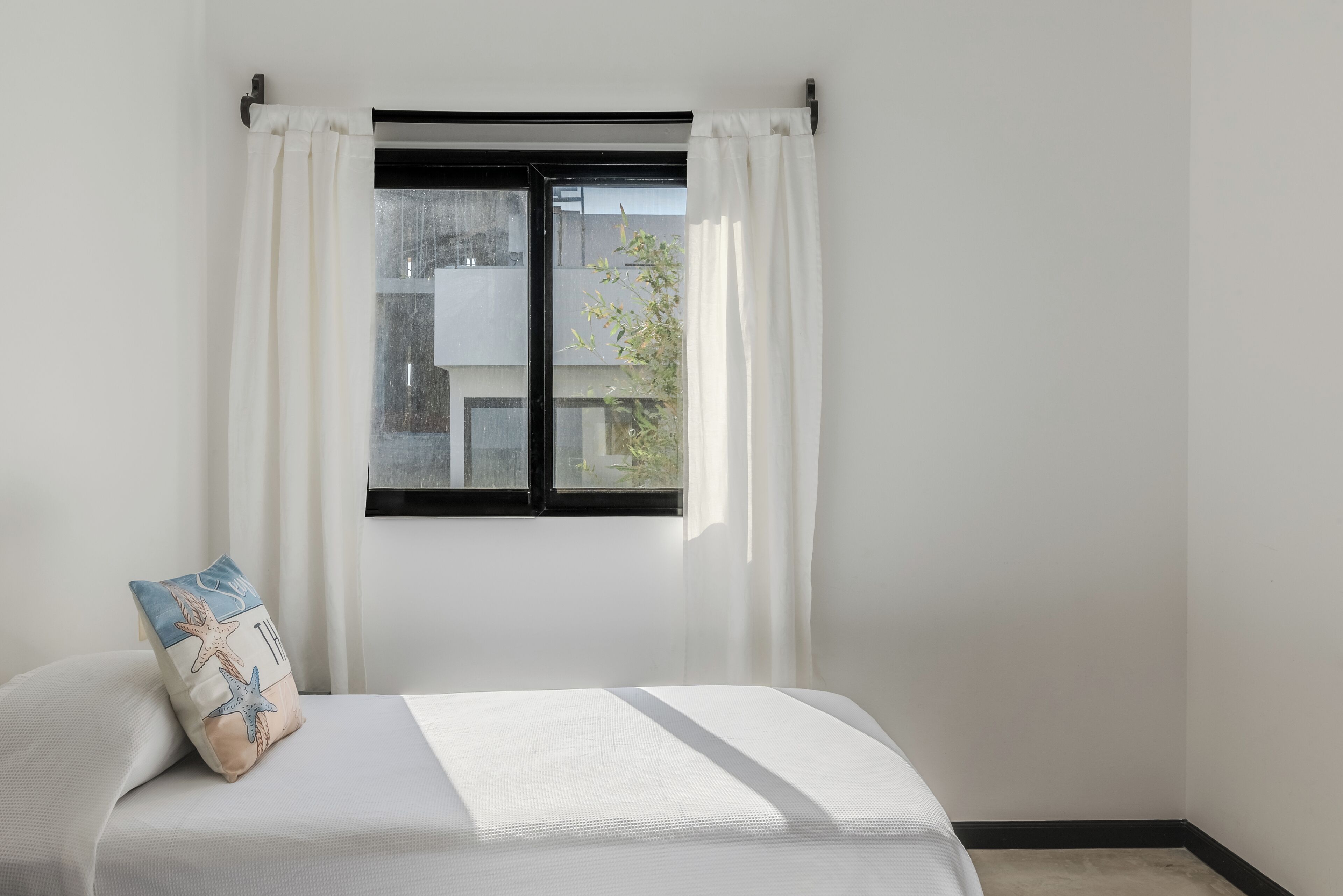 Un dormitorio con una cama, una ventana con cortinas blancas y vistas a edificios.
