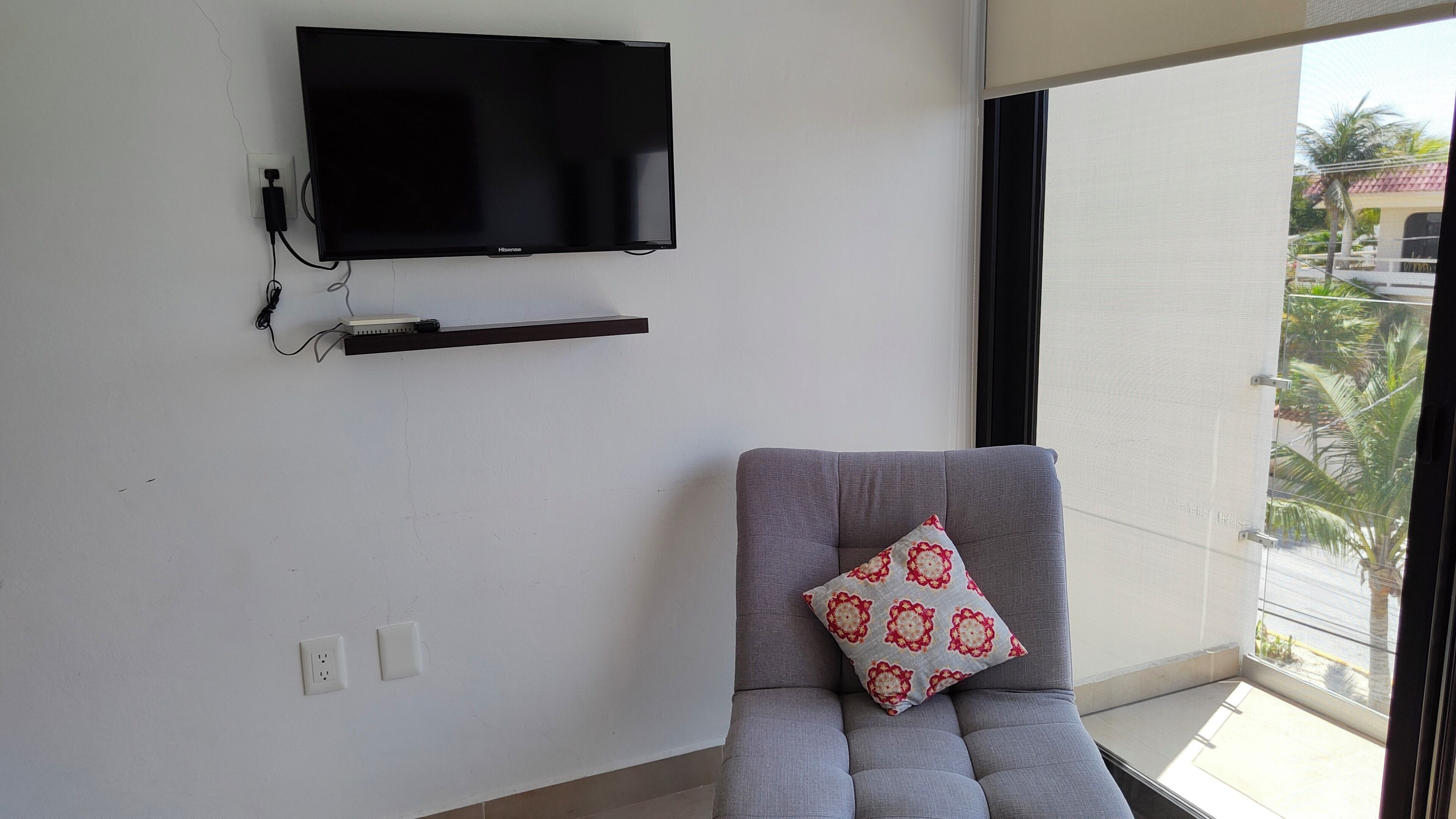 Ein Zimmer mit einem an der Wand montierten Flachbildfernseher, einem grauen Sofa mit einem gemusterten Kissen und einem Balkon mit Blick auf Palmen.