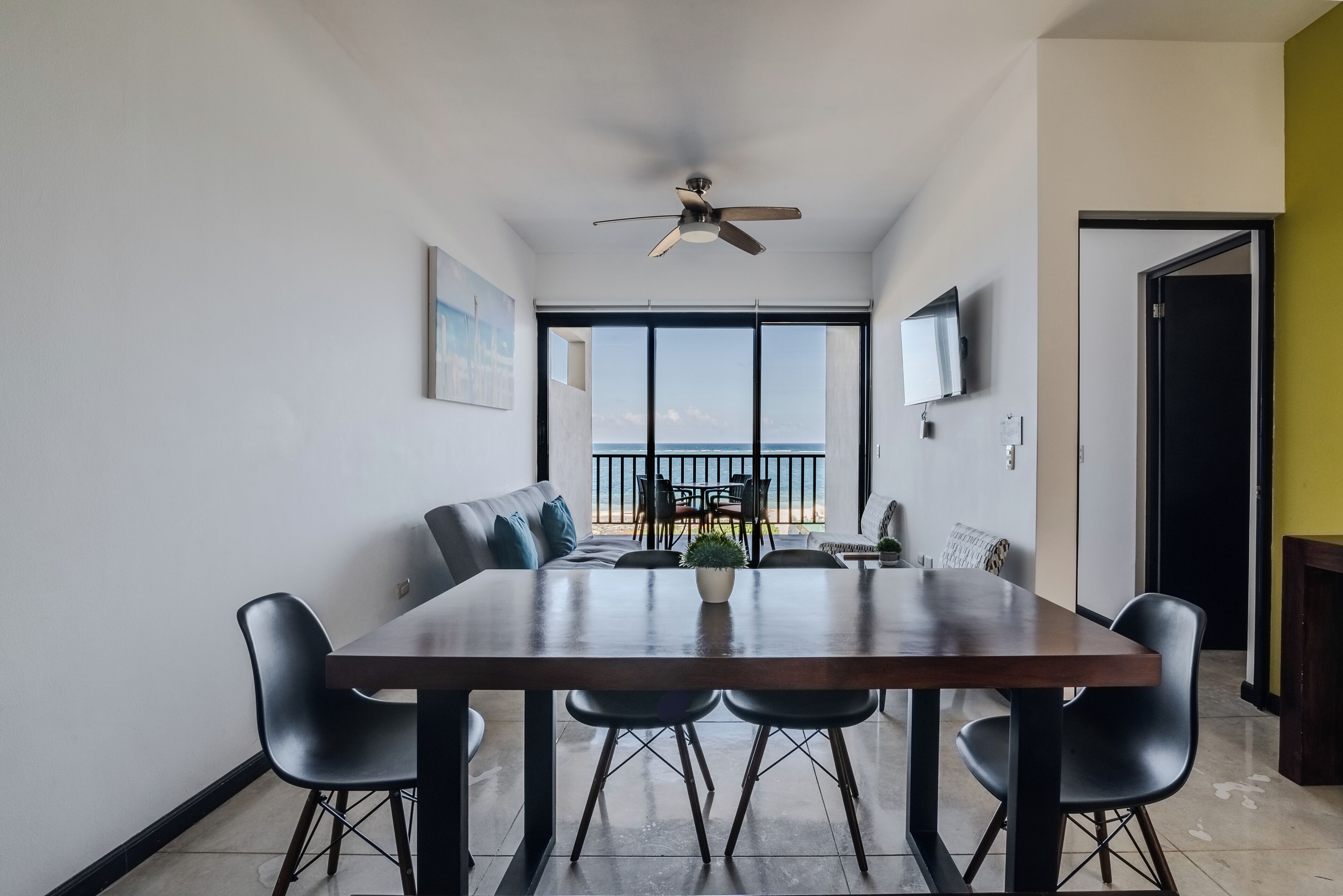 Zona pranzo con tavolo in legno, sedie nere, ventilatore a soffitto e vista sul mare attraverso ampie finestre.