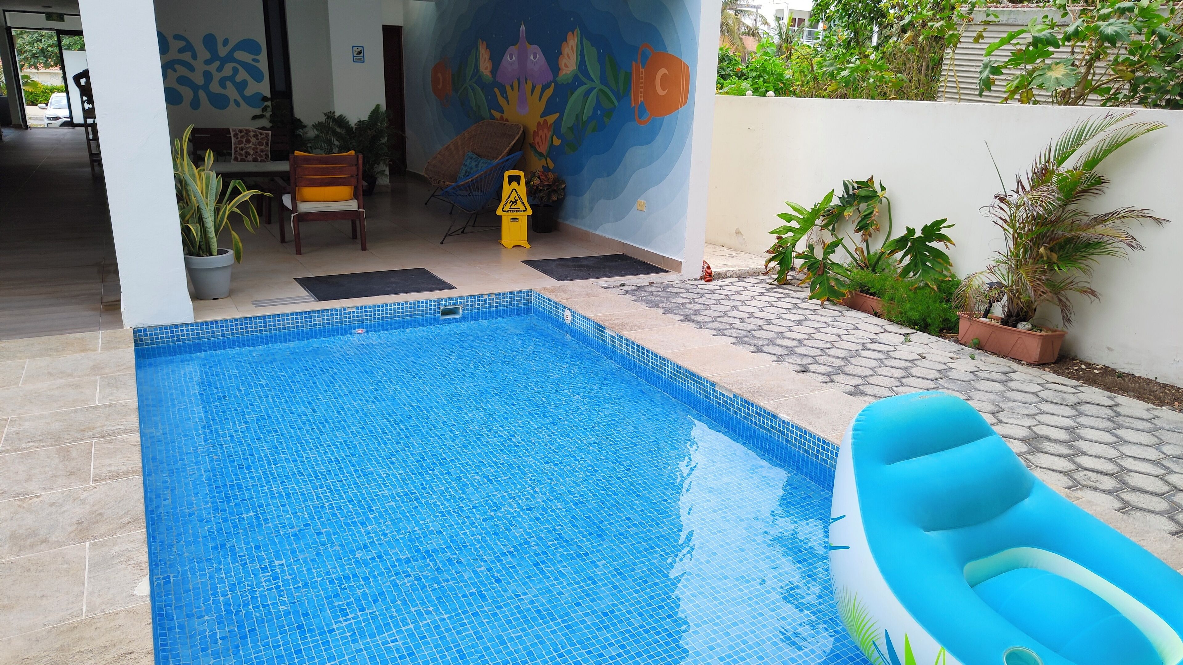 Une piscine dont le bord est recouvert de carreaux, entourée de plantes en pot et ornée d’une fresque murale.