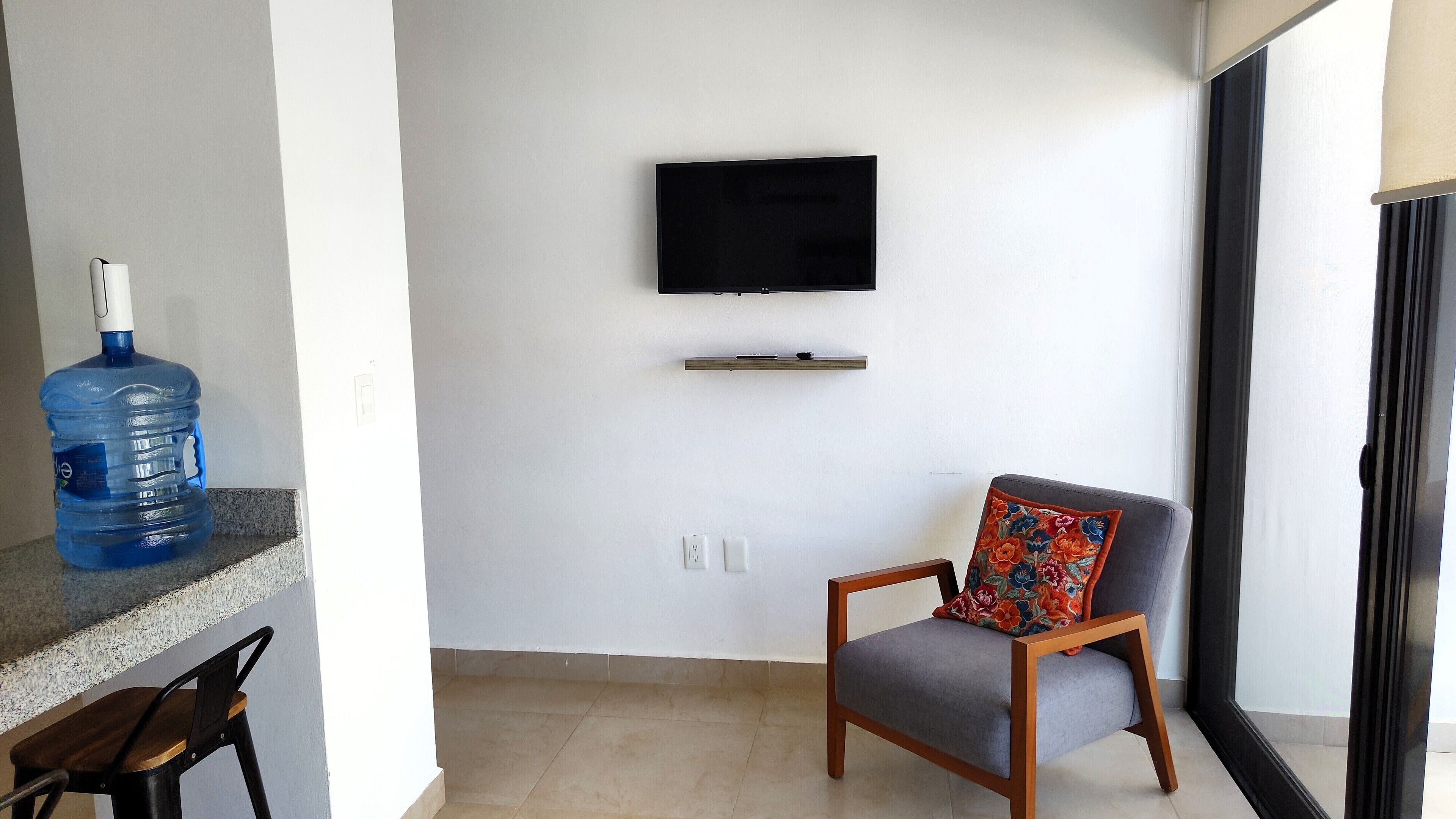 Un salon avec un téléviseur à écran plat fixé au mur, un grand distributeur d’eau, un fauteuil gris agrémenté d’un coussin à motifs floraux et une table à manger en bois.