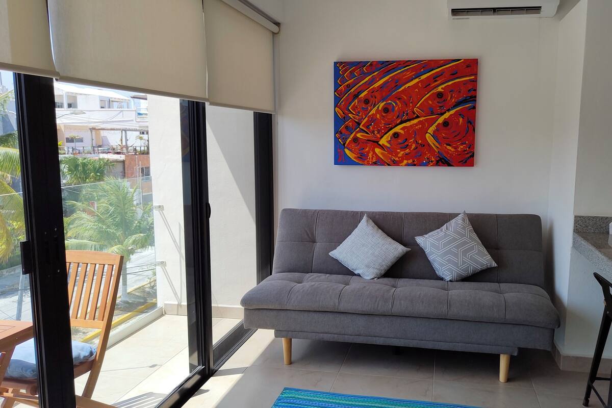 Una sala de estar moderna con un sofá gris, dos cojines, un cuadro abstracto de colores y vistas al exterior.