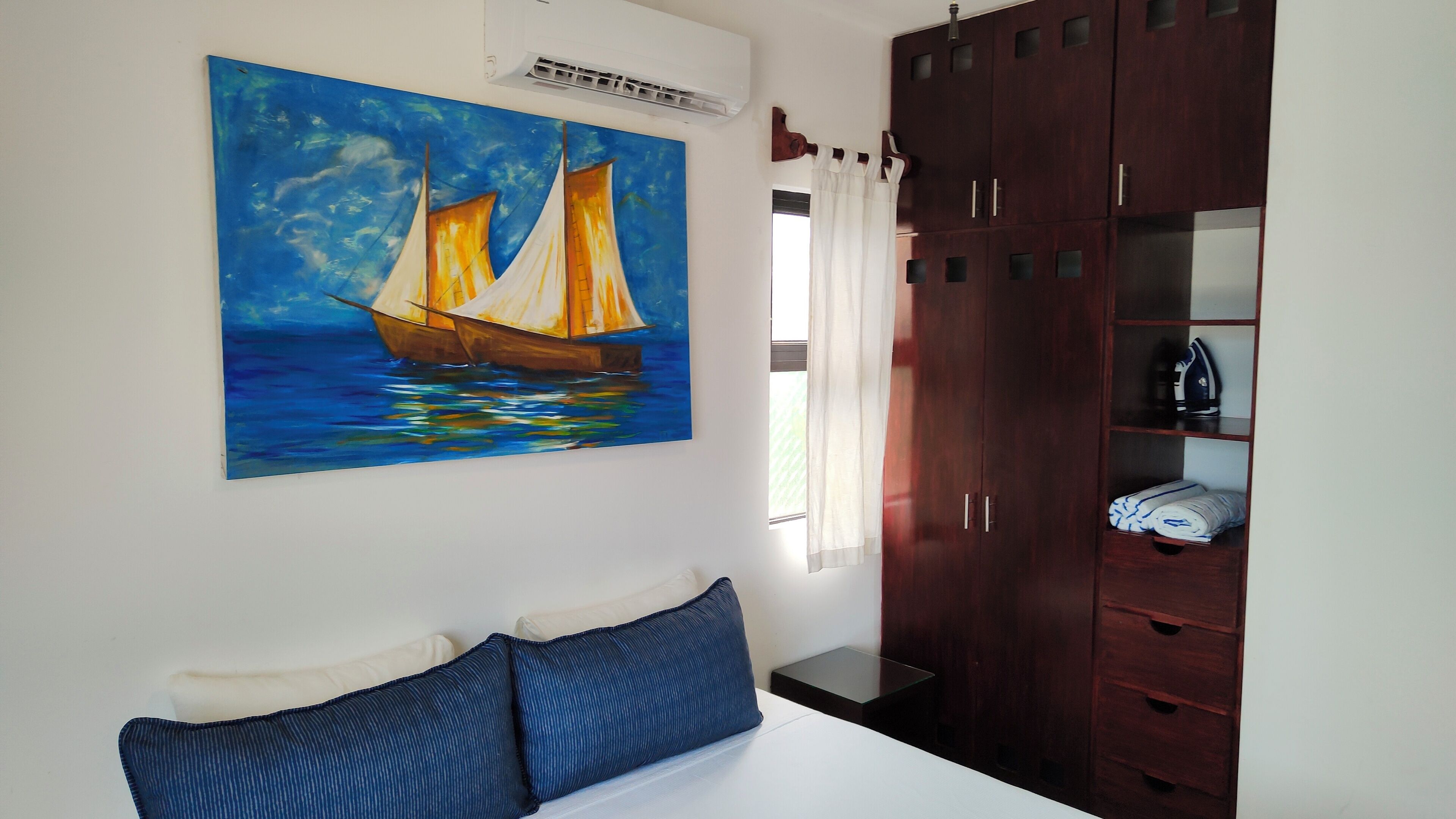 A bedroom with a bed, a painting of a sailboat, a wardrobe, and a window with curtains.