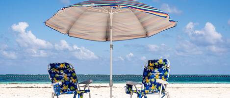 Two beach chairs under a striped umbrella on a sandy beach with the ocean in the background.