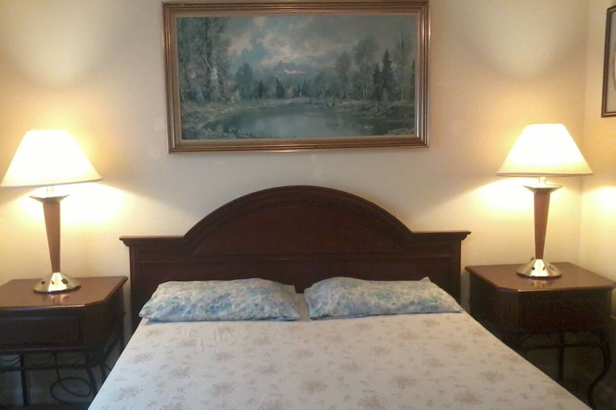 A bed with a floral bedspread, two bedside lamps, and a painting of a forest scene on the wall.