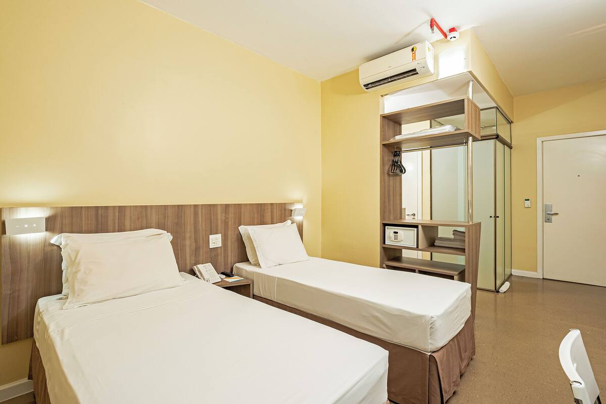Une chambre d’hôtel avec deux lits simples, une table de chevet avec un téléphone, un miroir, une armoire et un appareil de climatisation.
