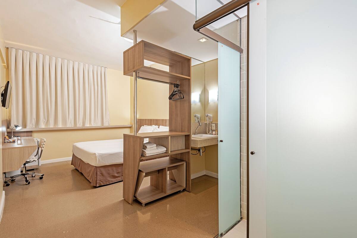 Une chambre d’hôtel comprenant un lit, un bureau, une chaise, un miroir et une salle de bain avec un lavabo et des toilettes.