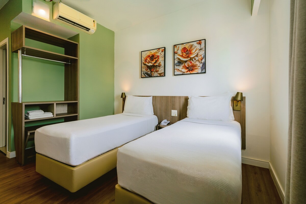 A hotel room with two single beds, a wooden wardrobe, and two floral paintings on the wall.